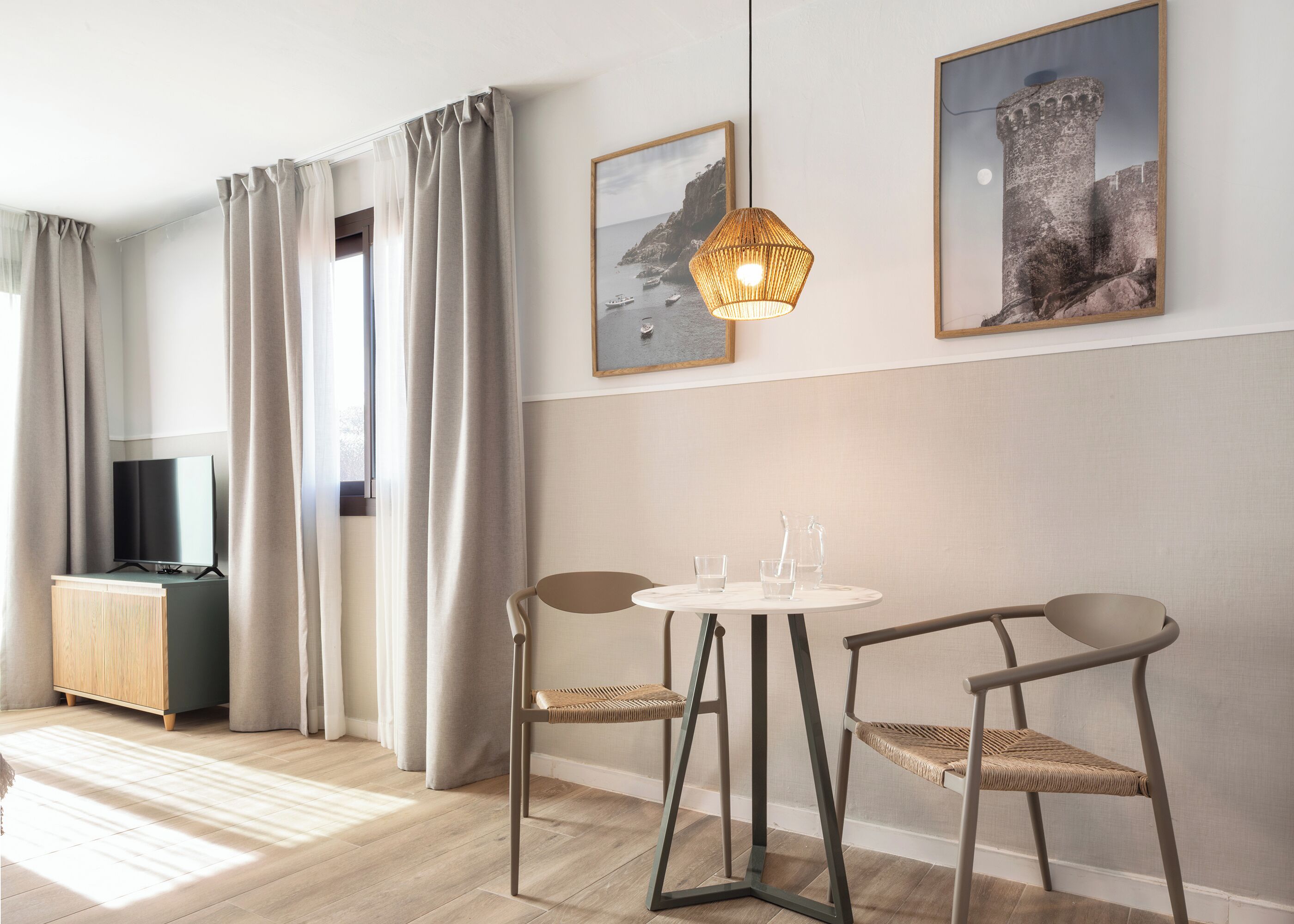 Habitació moderna amb televisió, dues cadires, una taula amb gots i una gerra, i dos quadres emmarcats a la paret.
