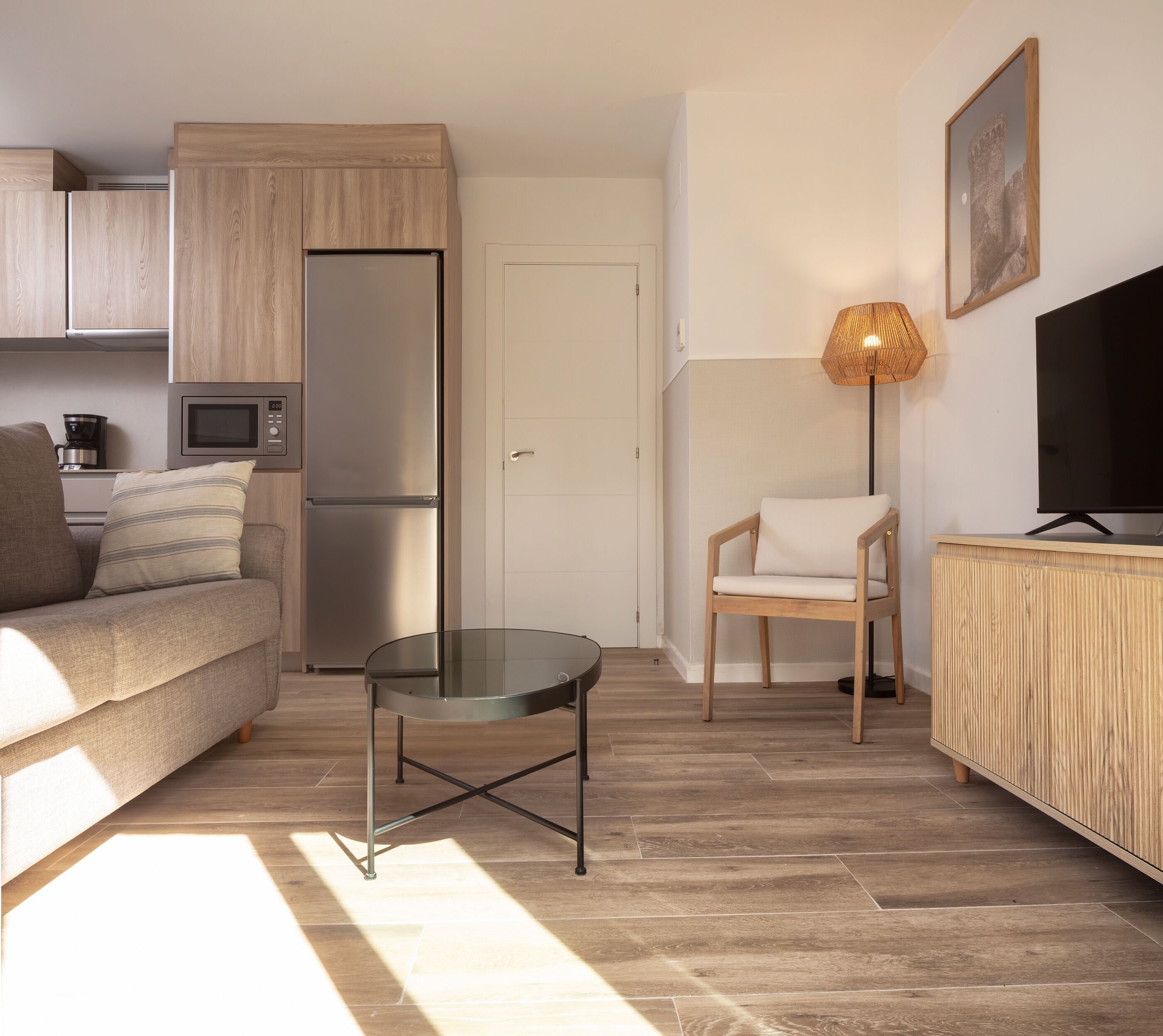 Ein kompakter Wohnraum mit einer Couch, einem Glastisch, einem Sessel, einem Fernseher und einer Küchenzeile mit Kühlschrank und Mikrowelle.