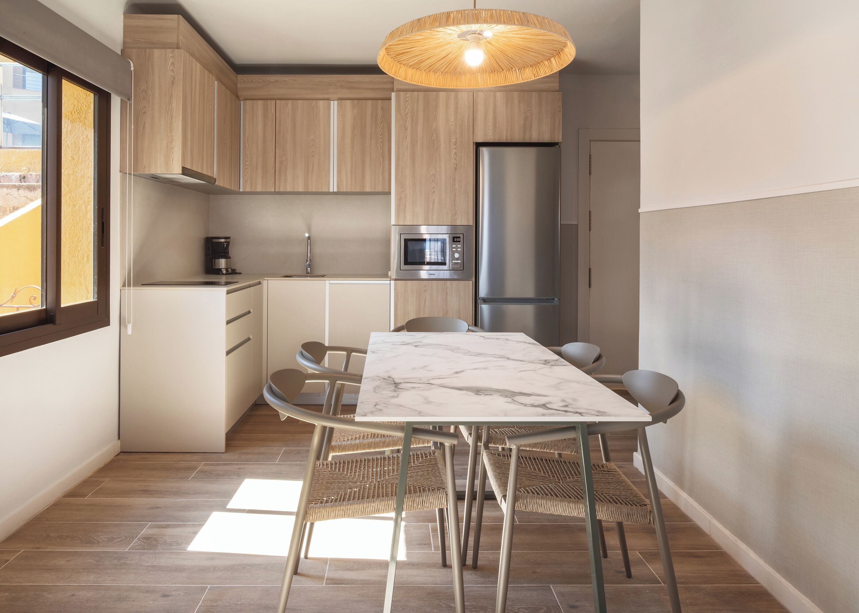 A modern kitchen with wooden cabinets, a stainless steel refrigerator, a microwave, and a dining area with a marble table and wicker chairs.