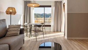 Ein modernes Hotelzimmer mit Sofa, Esstisch, Stühlen, einer Pendelleuchte und einem großen Fenster mit Vorhängen.