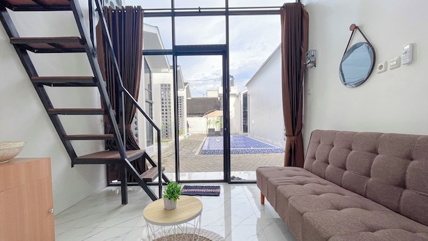A modern living room with a staircase, a sofa, a small table with a plant, and a view of the outside. - Sovilla II Yogyakarta (Depok)