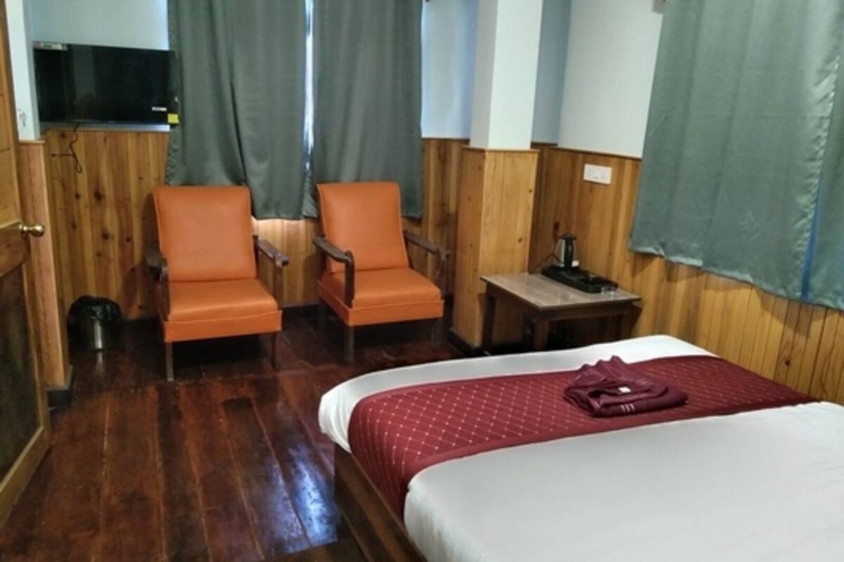 Habitación de hotel con piso de madera, dos sillas naranjas, una cama con colcha roja y blanca, una mesita con lámpara, y un televisor montado en la pared.