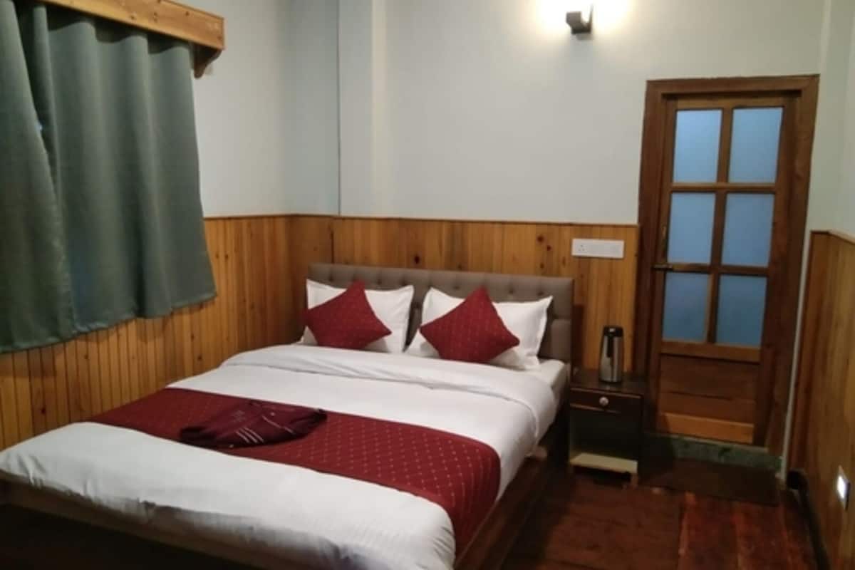 Un dormitorio con cabecera de madera, cama con ropa de cama blanca y almohadas rojas, mesita de noche con una lámpara y una puerta de madera.
