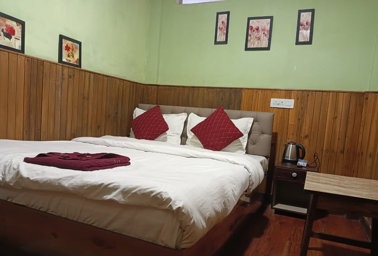 A hotel room with a wooden paneled wall, a bed with white bedding and red pillows, a small table with a kettle, and framed floral artwork on the wall.