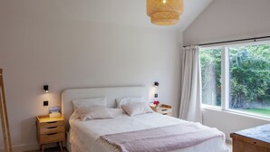 Un dormitorio con una cama grande, mesitas de noche, un aparador de madera, una lámpara colgante y una ventana con cortinas.