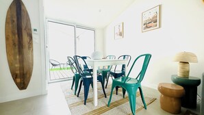 A dining area with a white table and teal chairs, a surfboard on the wall, and a view through a glass door to an outdoor space. - Maison jala (La Rochelle)