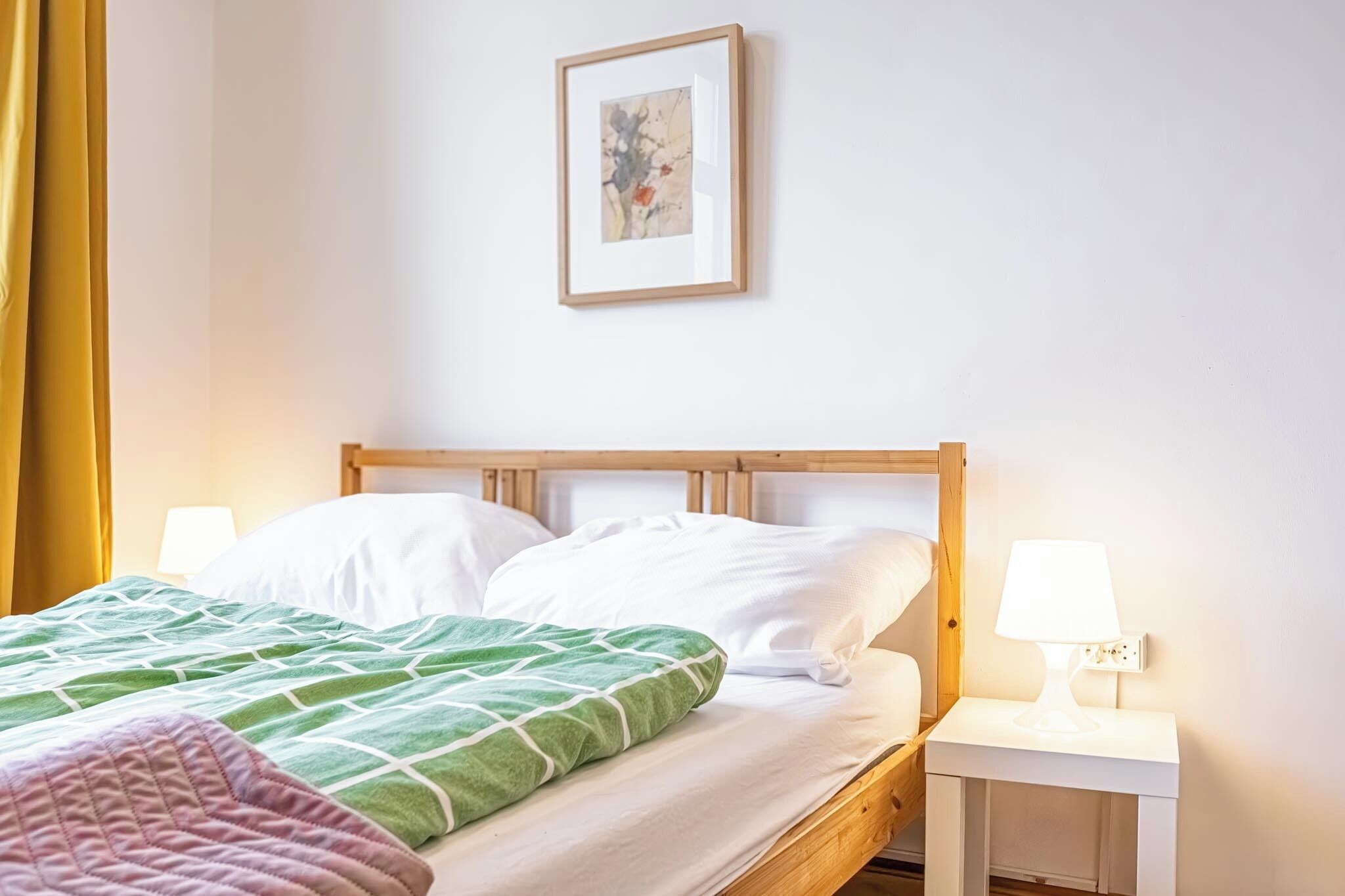 Ein ordentlich bezogenes Bett mit einer grünen und rosa Decke, einem Holzkopfstück und einer Nachttischlampe auf einem weißen Nachttisch.