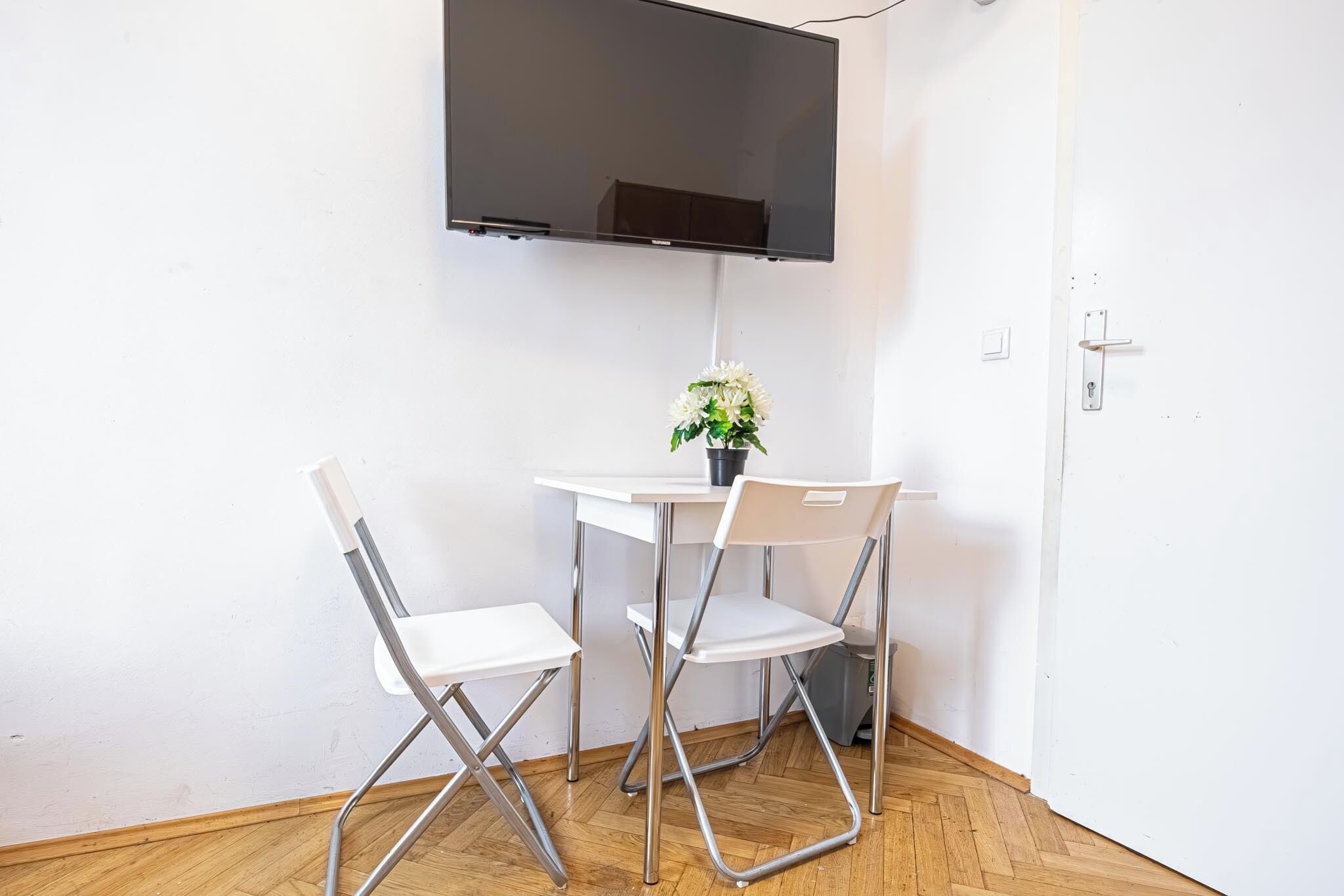 A small room with a white table and two white chairs, a potted plant, and a flat-screen TV mounted on the wall.