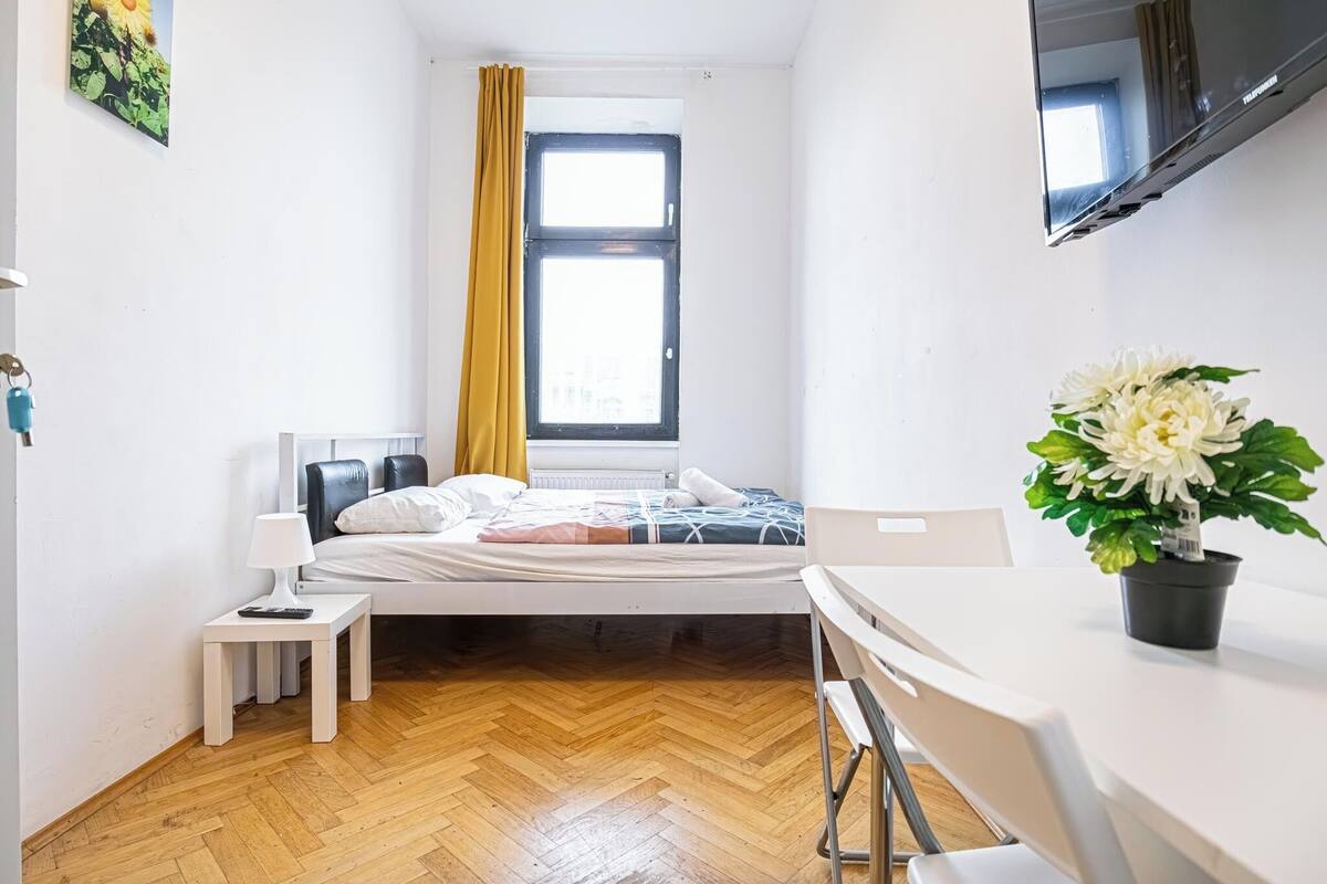 A small, clean room with a bed, a small table, a chair, a potted plant, and a TV mounted on the wall.