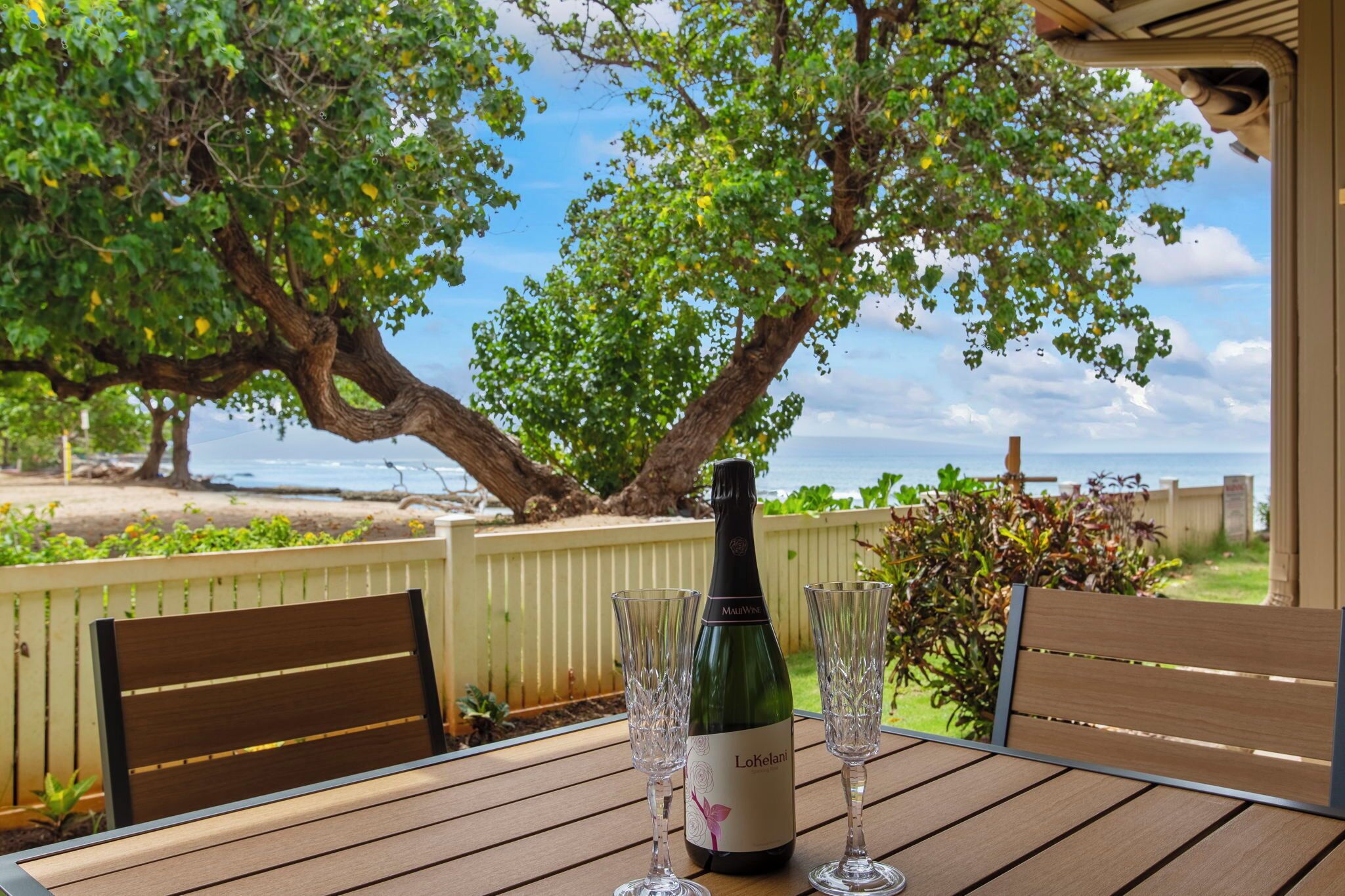 A table with a bottle of wine and two glasses on a wooden deck overlooking a beach.
