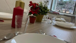A dining table with a white plate, cutlery, and a red candle. There are two potted plants, one with red flowers and another with green leaves. A window with a view of trees is visible in the background.
