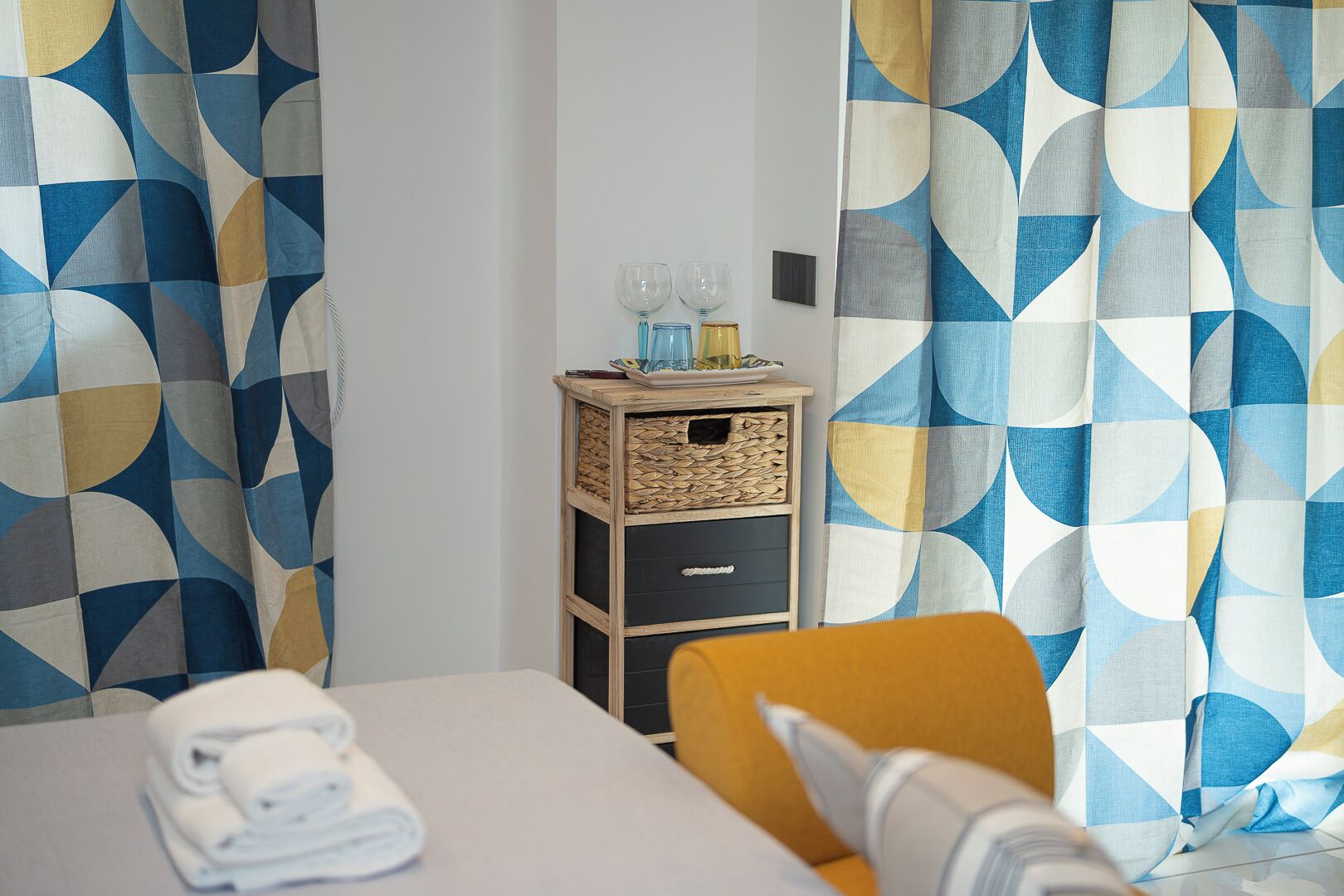 A hotel room with a bed, a small wooden side table with a woven basket, a chair, and a table with a rolled towel.