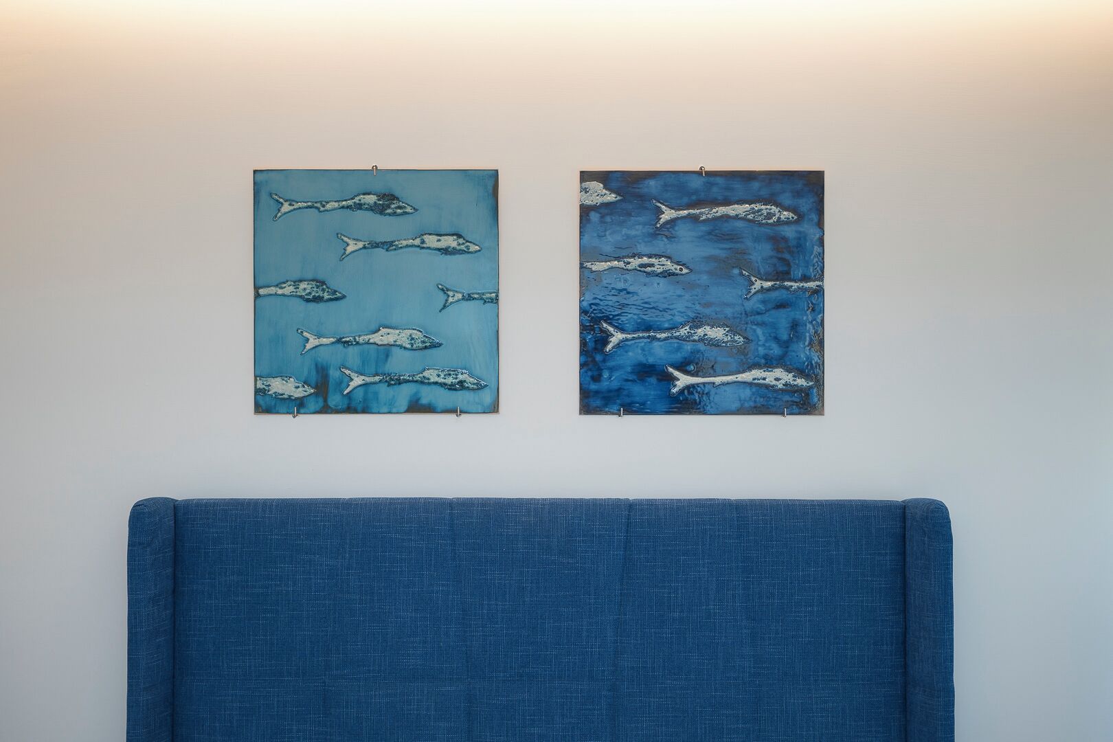 Dos obras de arte colgadas en la pared, que representan peces, sobre un cabecero tapizado en azul.