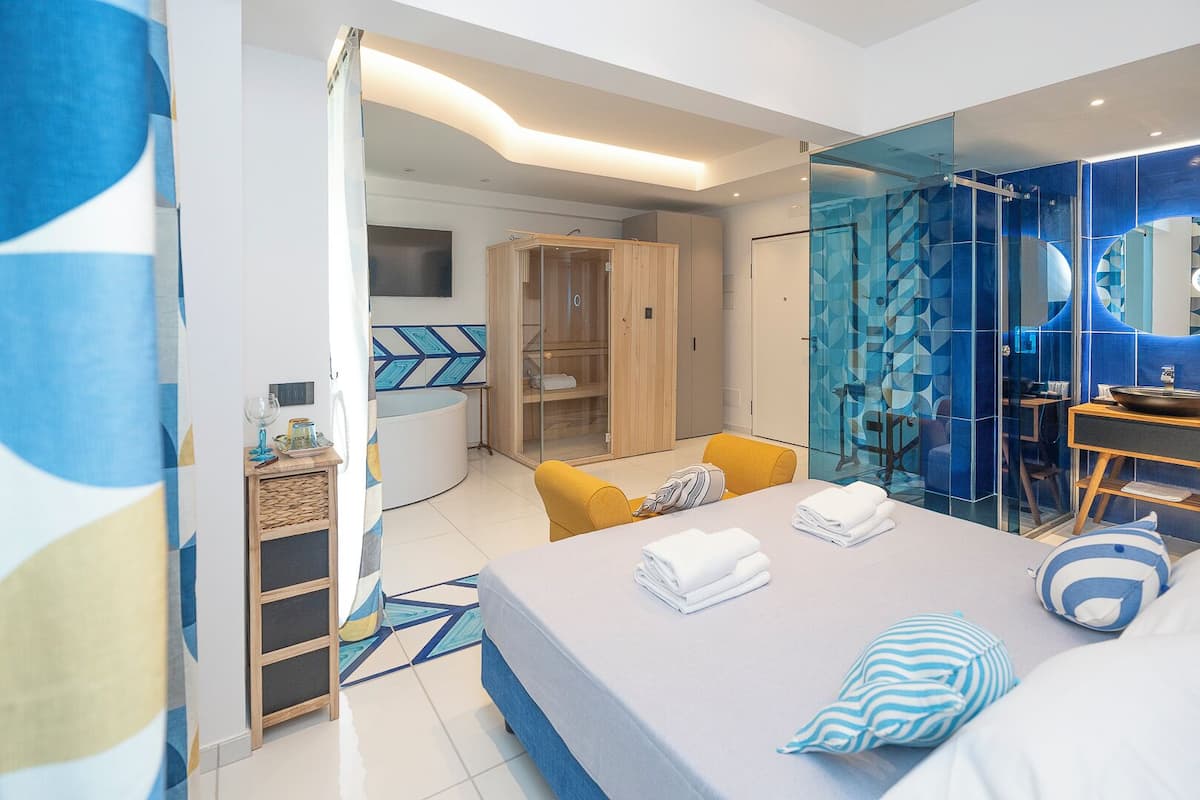 A modern hotel room with a bed, a seating area with yellow chairs, a glass-enclosed shower area, and a wooden wardrobe.
