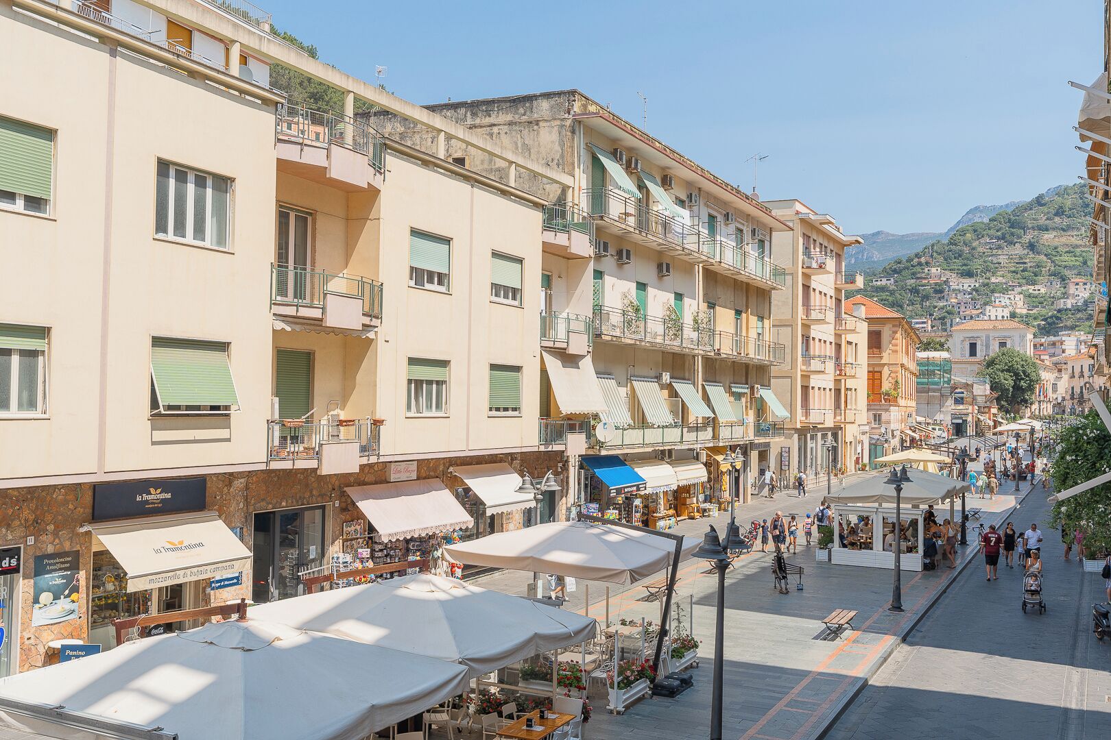 Una calle animada con edificios de varios pisos, cafés al aire libre y una vista despejada de la colina circundante.