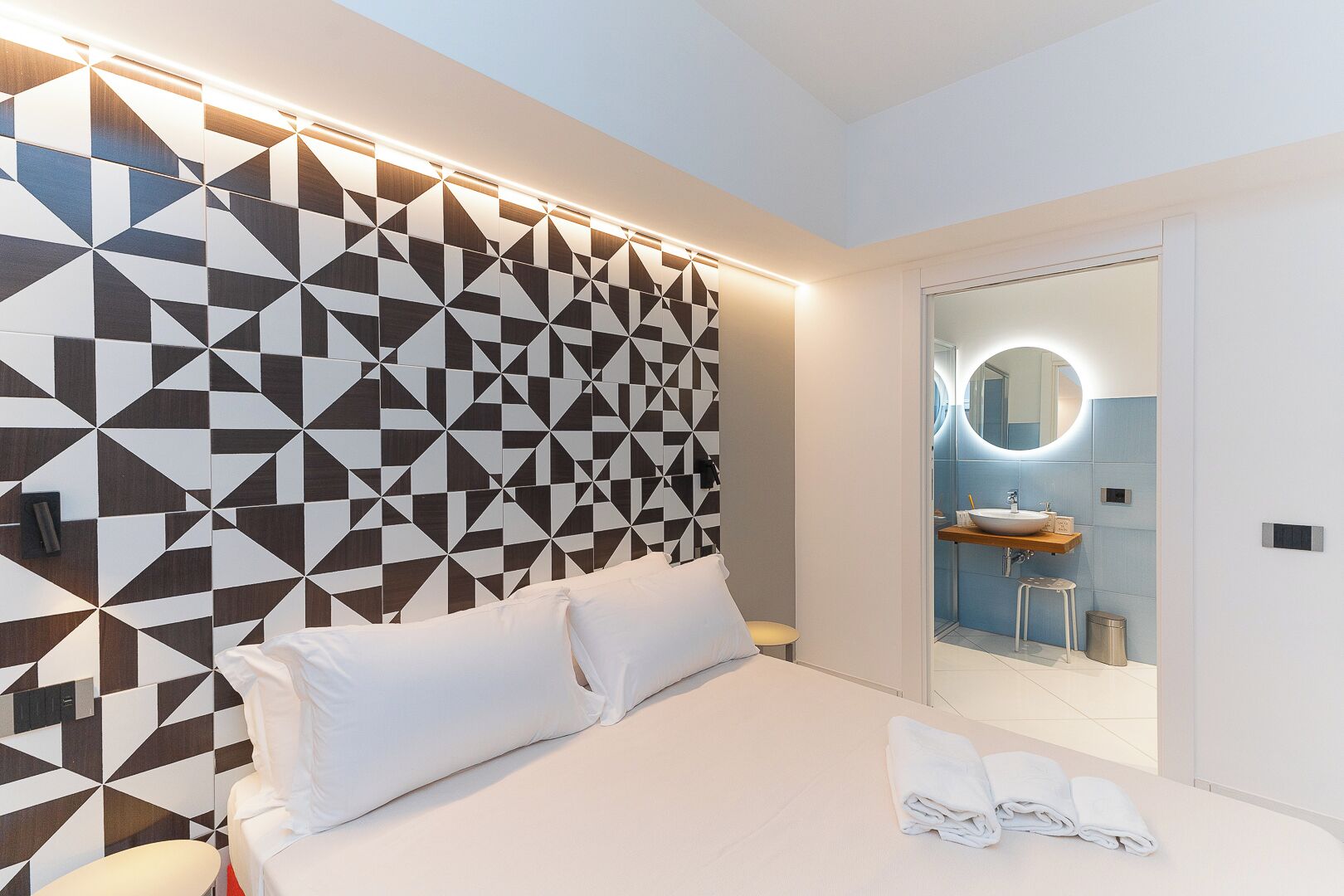 Quarto de hotel moderno com cabeceira estampada em padrão geométrico, uma cama grande com roupa de cama branca, um espelho redondo sobre a pia e um banquinho pequeno.