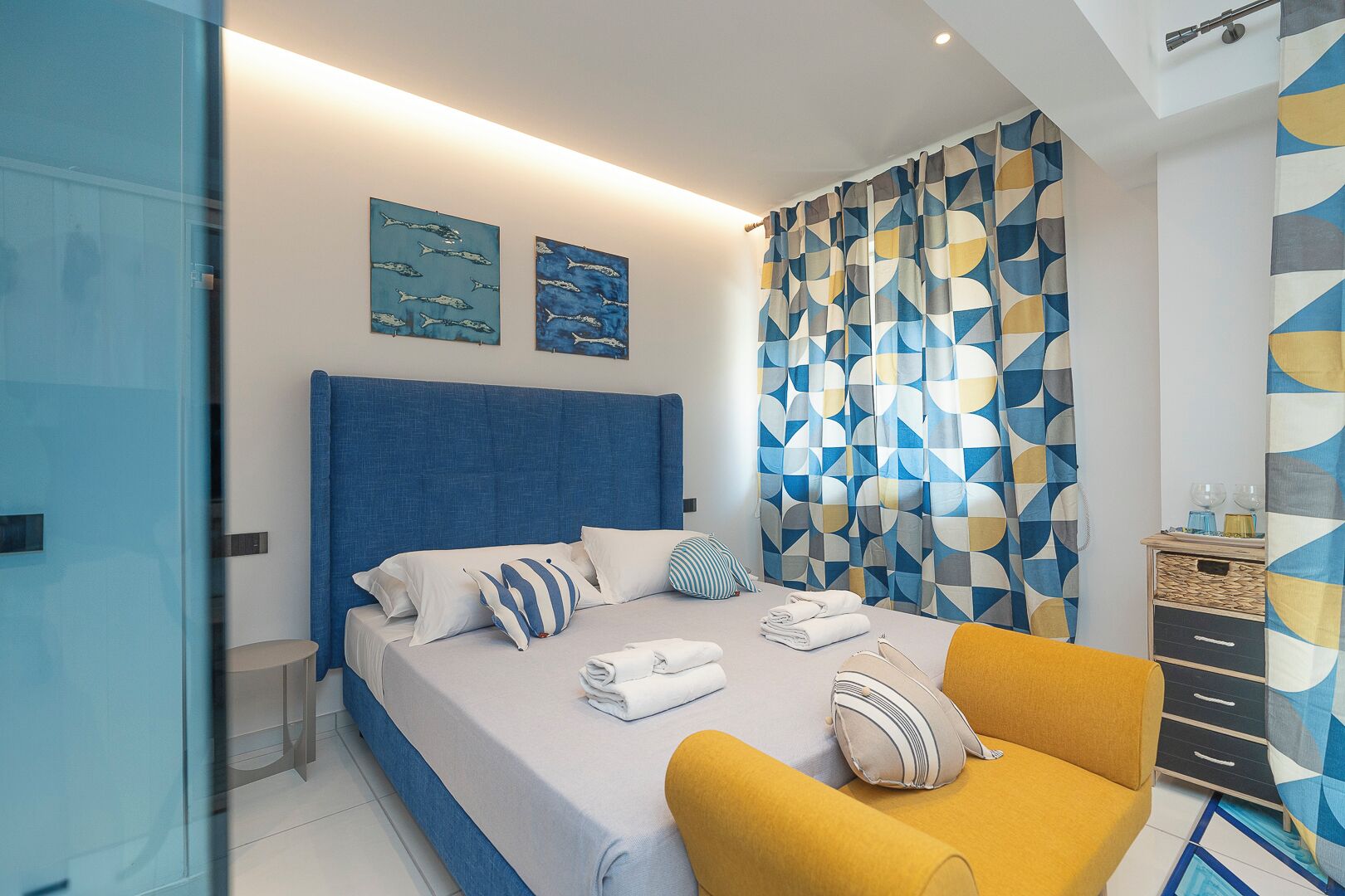 A modern bedroom with a large bed, a yellow armchair, and geometric-patterned curtains.