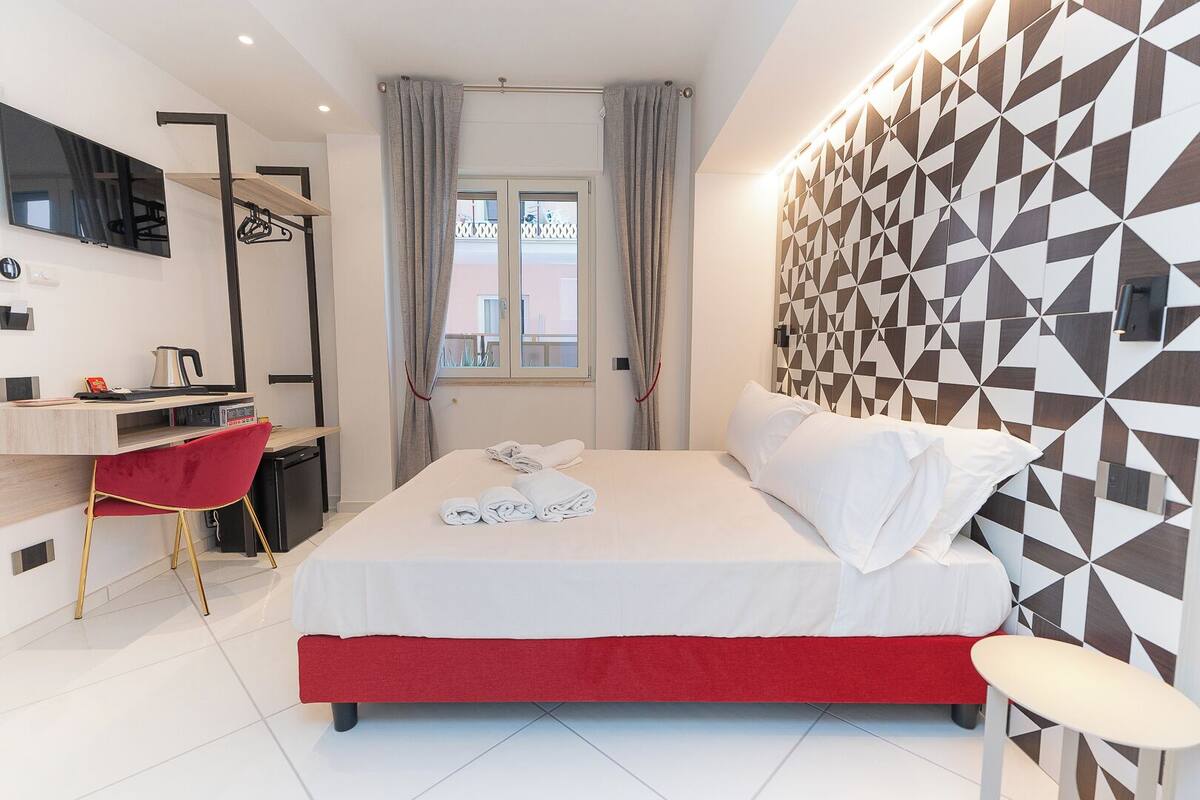 A modern hotel room with a bed, a desk, a red chair, and a geometric-patterned wall.