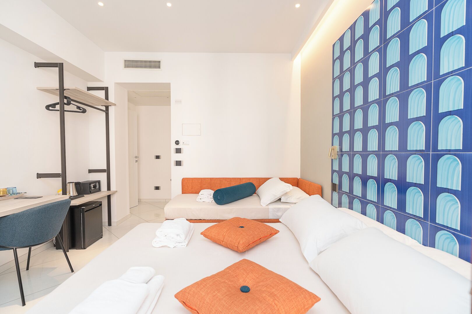 A modern hotel room with a bed, a desk, and a chair. There is a wall with a blue pattern and a door leading to another room.