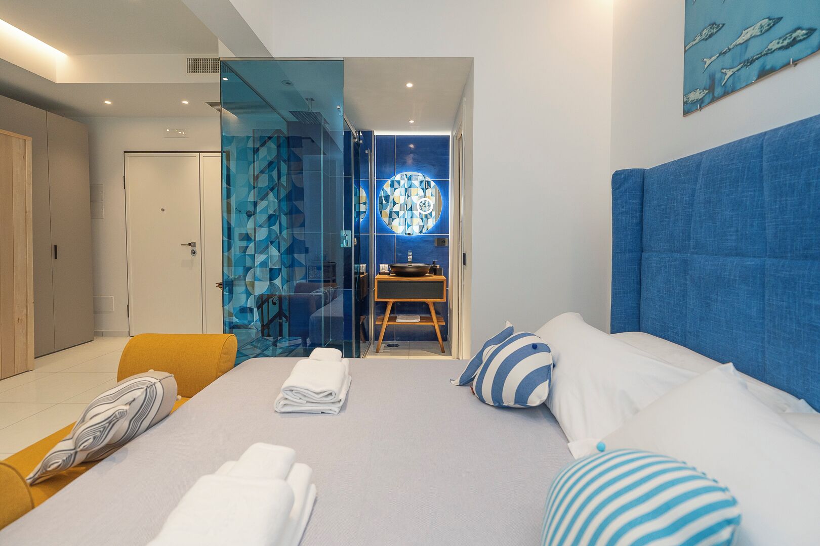 A modern hotel room with a bed, a yellow armchair, a glass partition, and a bathroom visible through the partition.