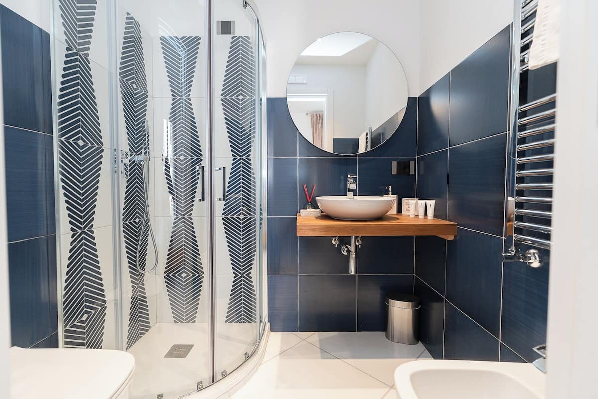 A modern bathroom with a glass shower enclosure, a round mirror, a wooden vanity, and a white sink.