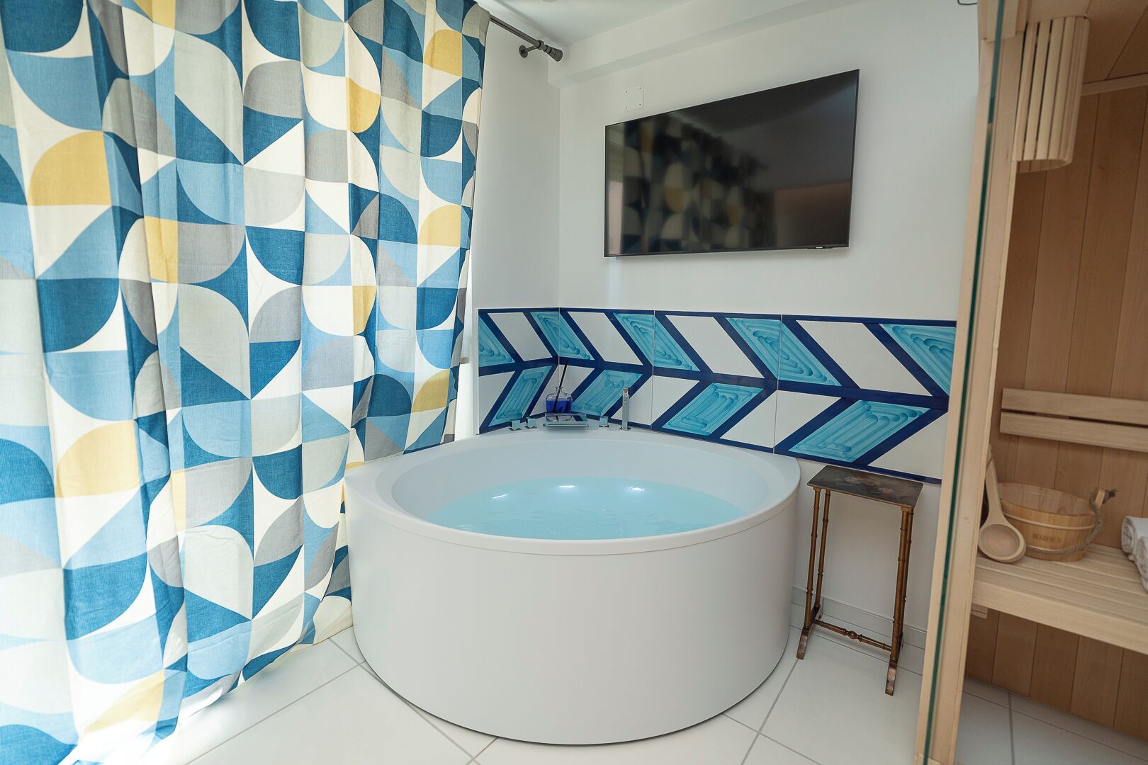 Ein Badezimmer mit freistehender Badewanne, einer Duschvorhang mit geometrischem Muster und einem an der Wand montierten Fernseher.