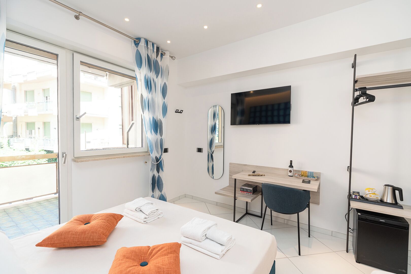 A modern hotel room with a bed, a desk, a chair, a TV, and a small kitchenette.