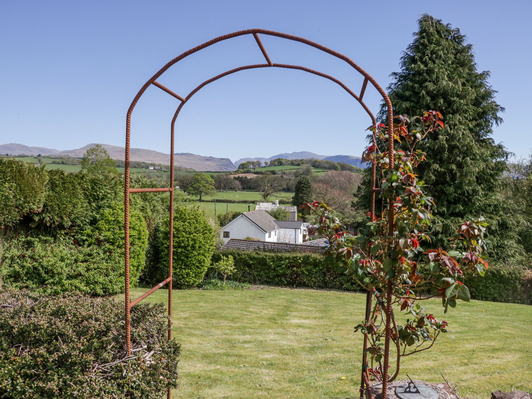 Un jardín con un arco de metal, abundante vegetación y una casa al fondo.