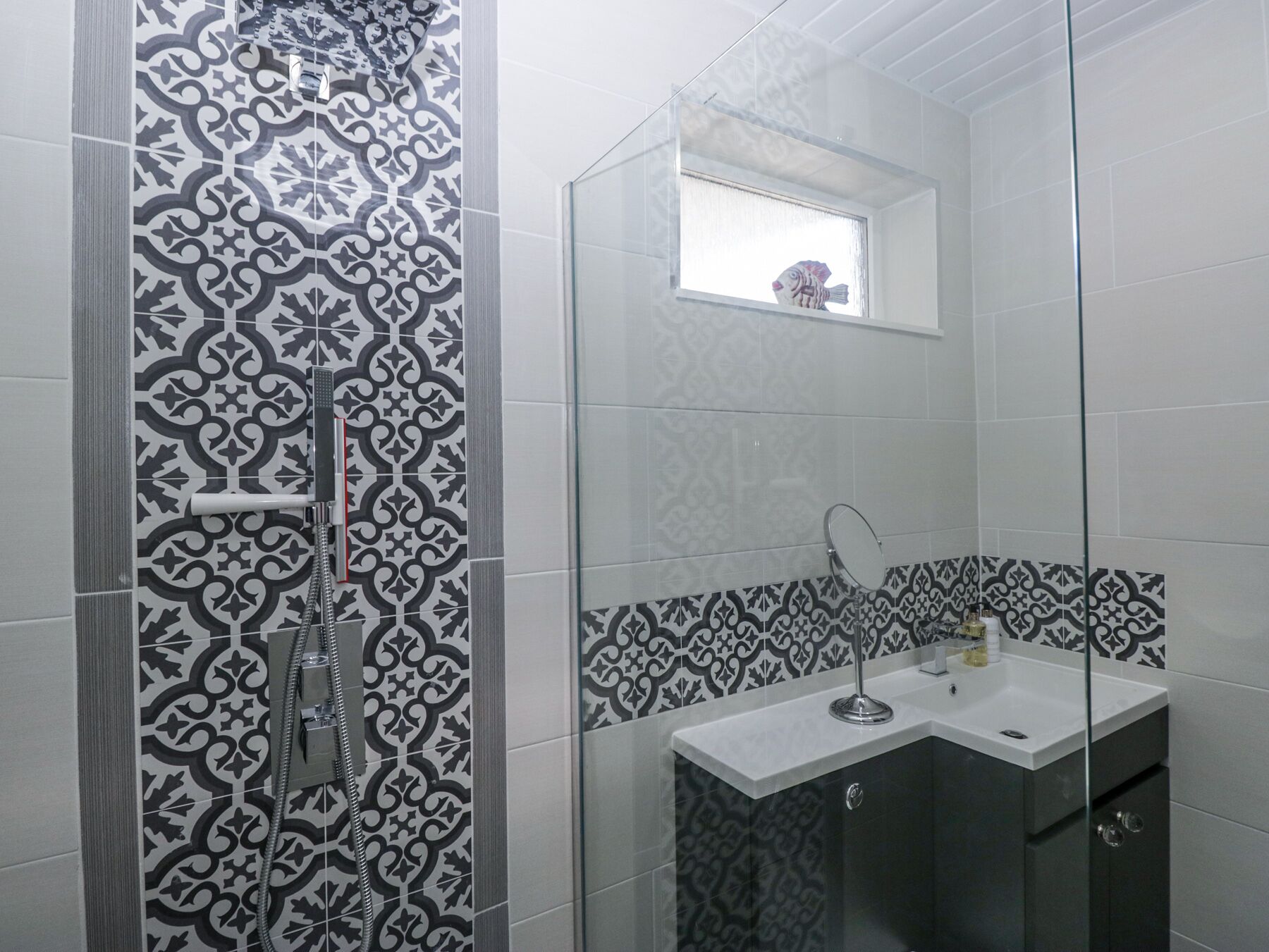A bathroom with patterned tiles, a glass shower enclosure, a sink with a mirror, and a small window above the mirror.