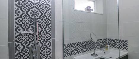 A bathroom with patterned tiles, a glass shower enclosure, a sink with a mirror, and a small window above the mirror.