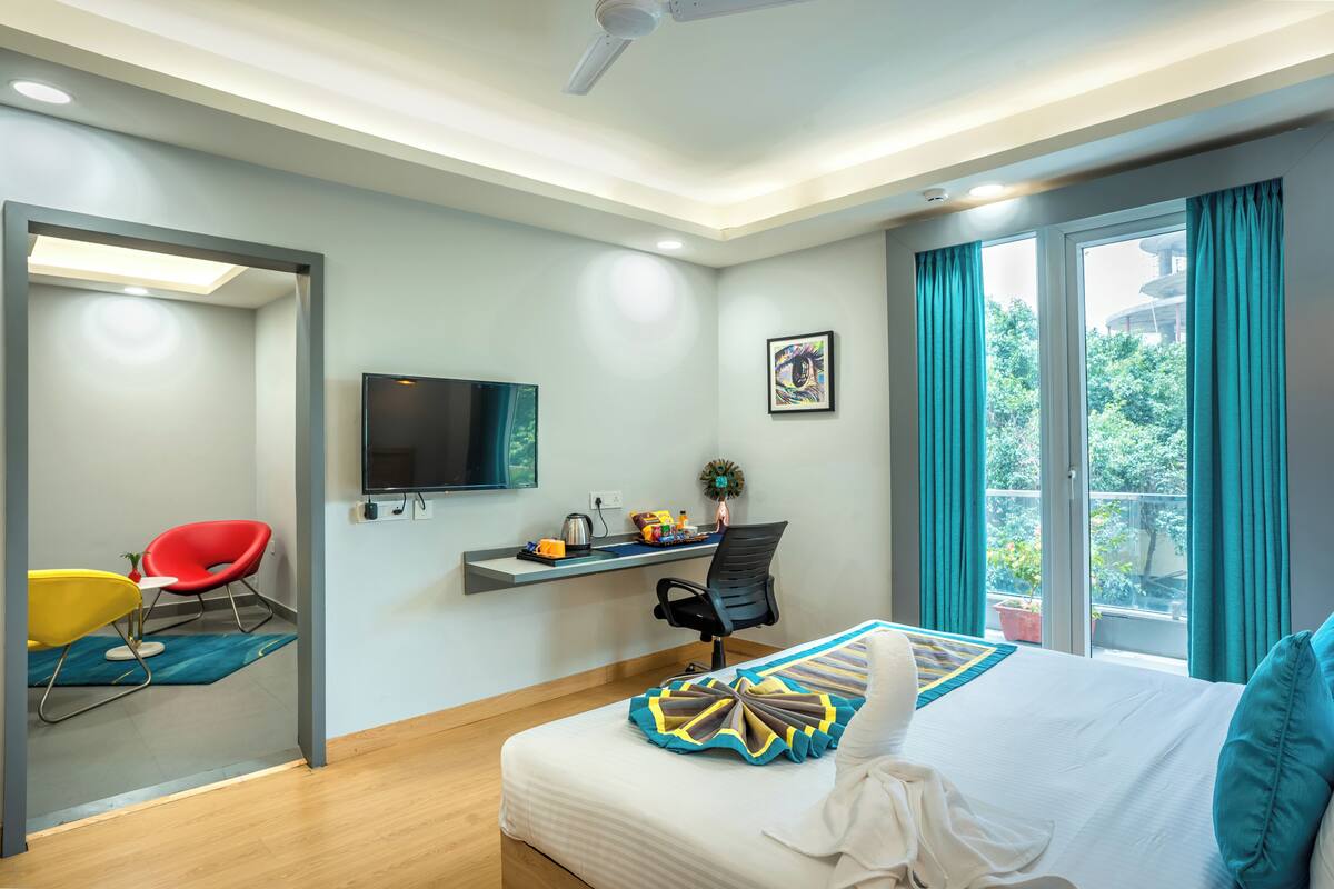 A modern hotel room with a bed, a desk, a chair, a TV, and a balcony with curtains.