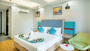 Una habitación de hotel con una cama, mesitas de noche, un sillón verde y una pequeña mesa con una planta.