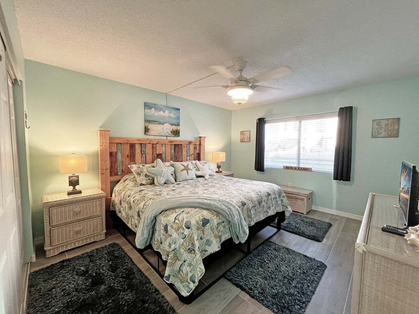 Un dormitorio con cama, mesitas de noche, ventilador de techo, ventana con cortinas y un cuadro en la pared.