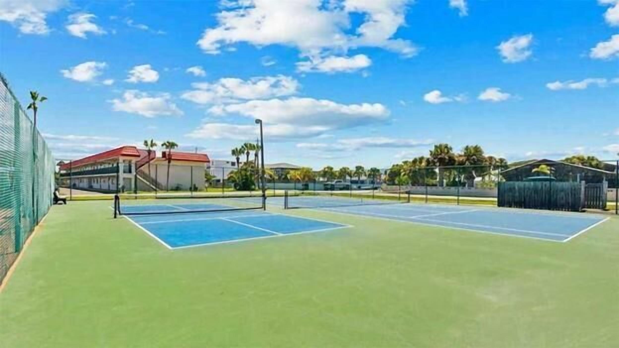 Cancha de tennis a l'aire lliure amb superfície verda, envoltada per una tanca i edificis al fons.