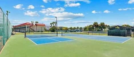 An outdoor tennis court with a green surface, surrounded by a fence, and buildings in the background.