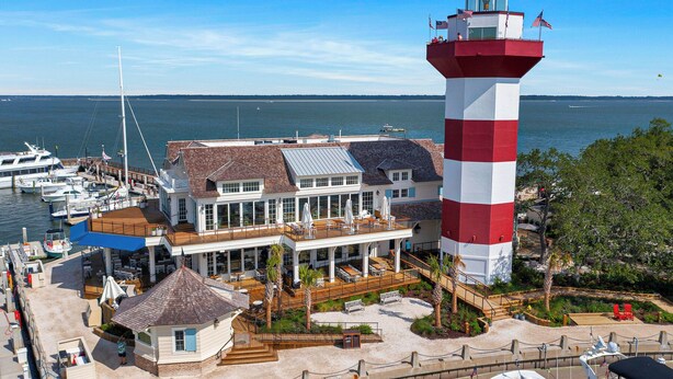 Un faro con una torre a rayas rojas y blancas, un edificio blanco con terraza de madera, y un puerto deportivo con barcos.