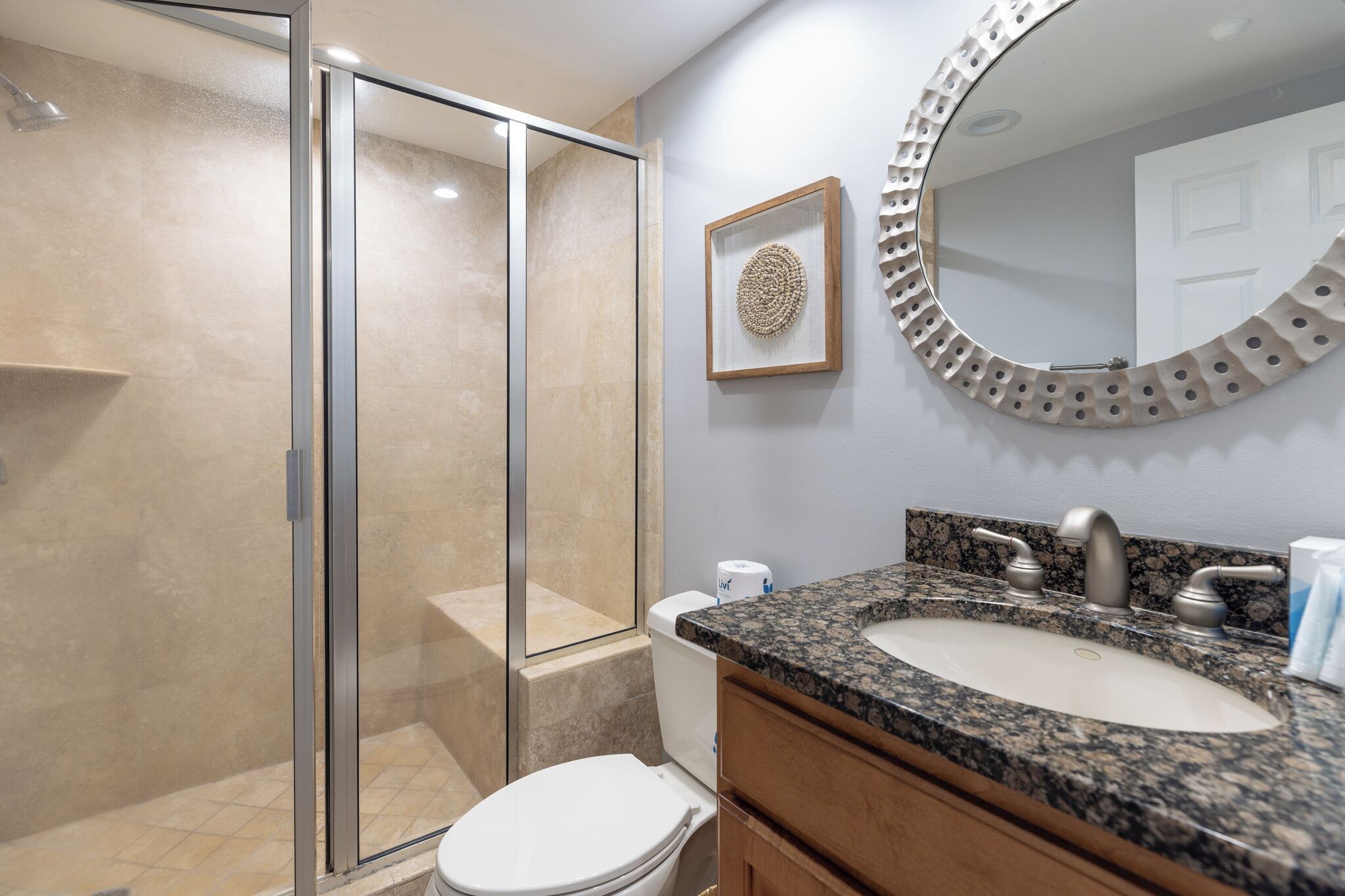 A bathroom with a glass shower enclosure, a toilet, a sink with a granite countertop, and a mirror with a decorative frame.
