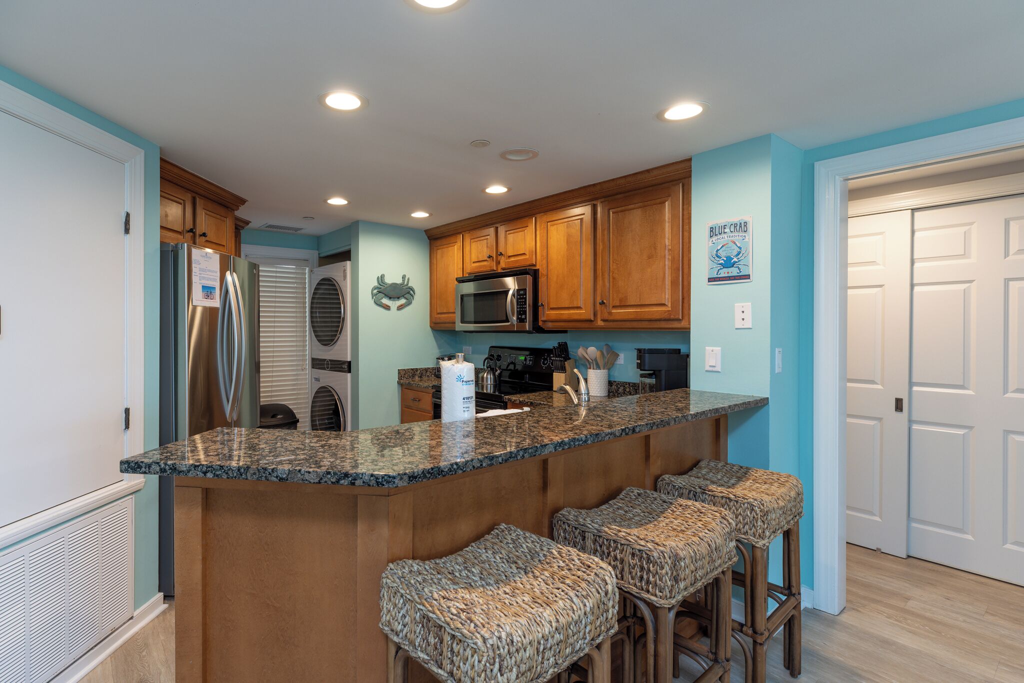 A kitchen with a granite countertop, wooden cabinets, a stainless steel refrigerator, a microwave, and a sink. There are three woven bar stools.