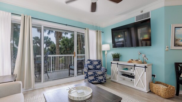 A living room with a sliding glass door, a ceiling fan, a TV mounted on the wall, a blue patterned chair, a wooden coffee table, and a white cabinet with shelves.