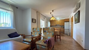 A living room with a sofa, two armchairs, a wooden table, and a kitchen area with wooden cabinets and a refrigerator. - Villa with private Pool/Aircon/Wi-Fi - Villa Montgrí (L'Escala)