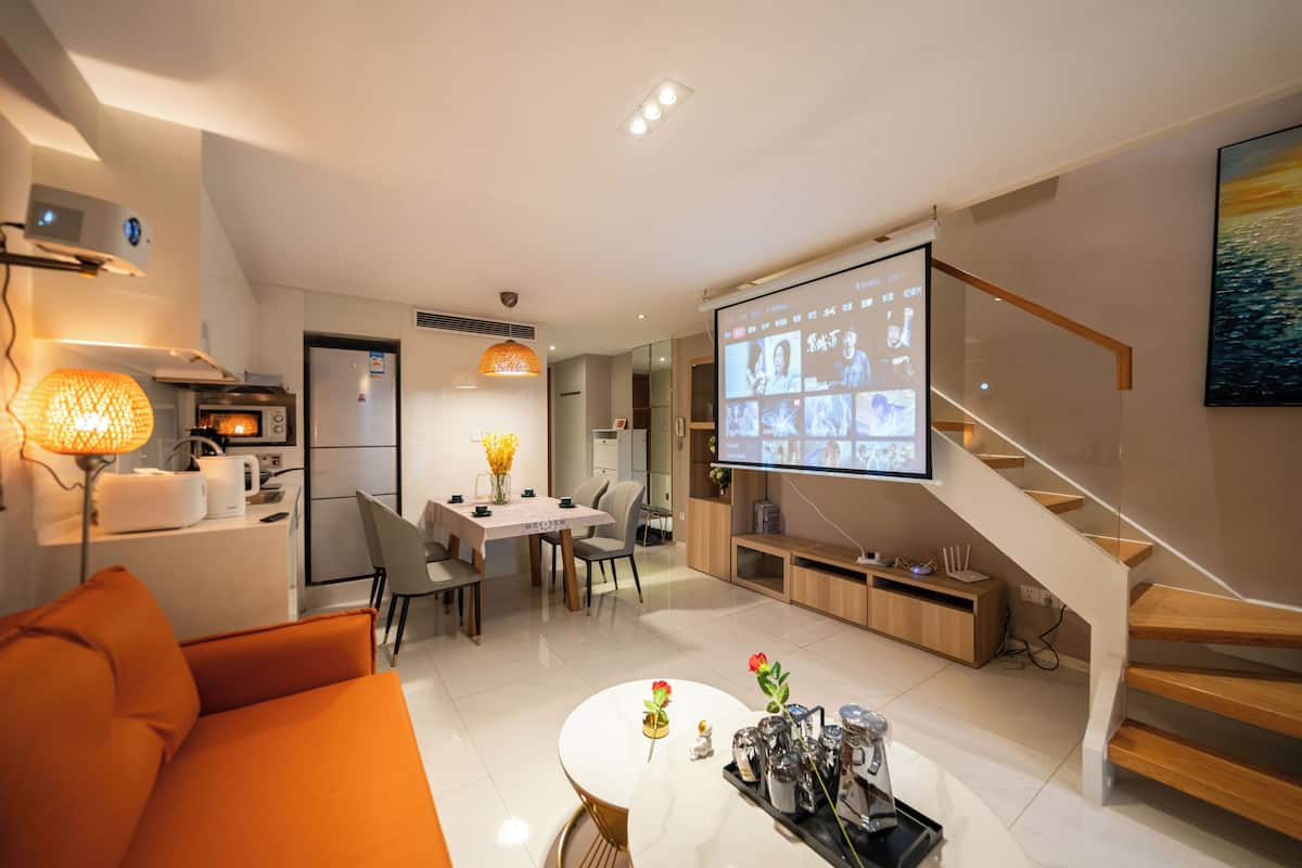 Una sala de estar moderna con un sofá, una mesa de centro y un televisor mostrando una videollamada. Se observa una escalera y una zona de cocina al fondo.