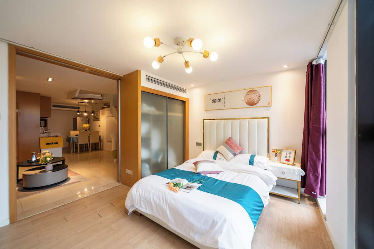 Habitación de hotel moderna con una cama grande, una mesita de noche con lámpara, un armario y vista al comedor.
