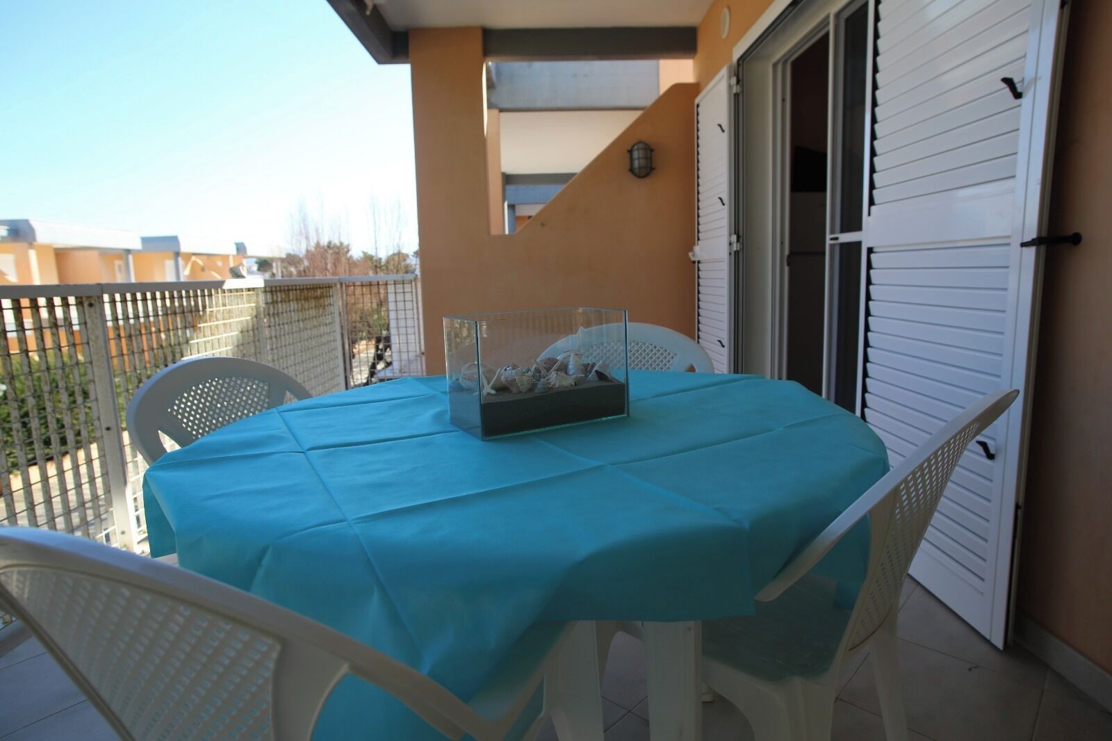 Een balkon met een tafel, bedekt met een blauwe tafelkleed, witte stoelen en een glazen displaykast met spullen.