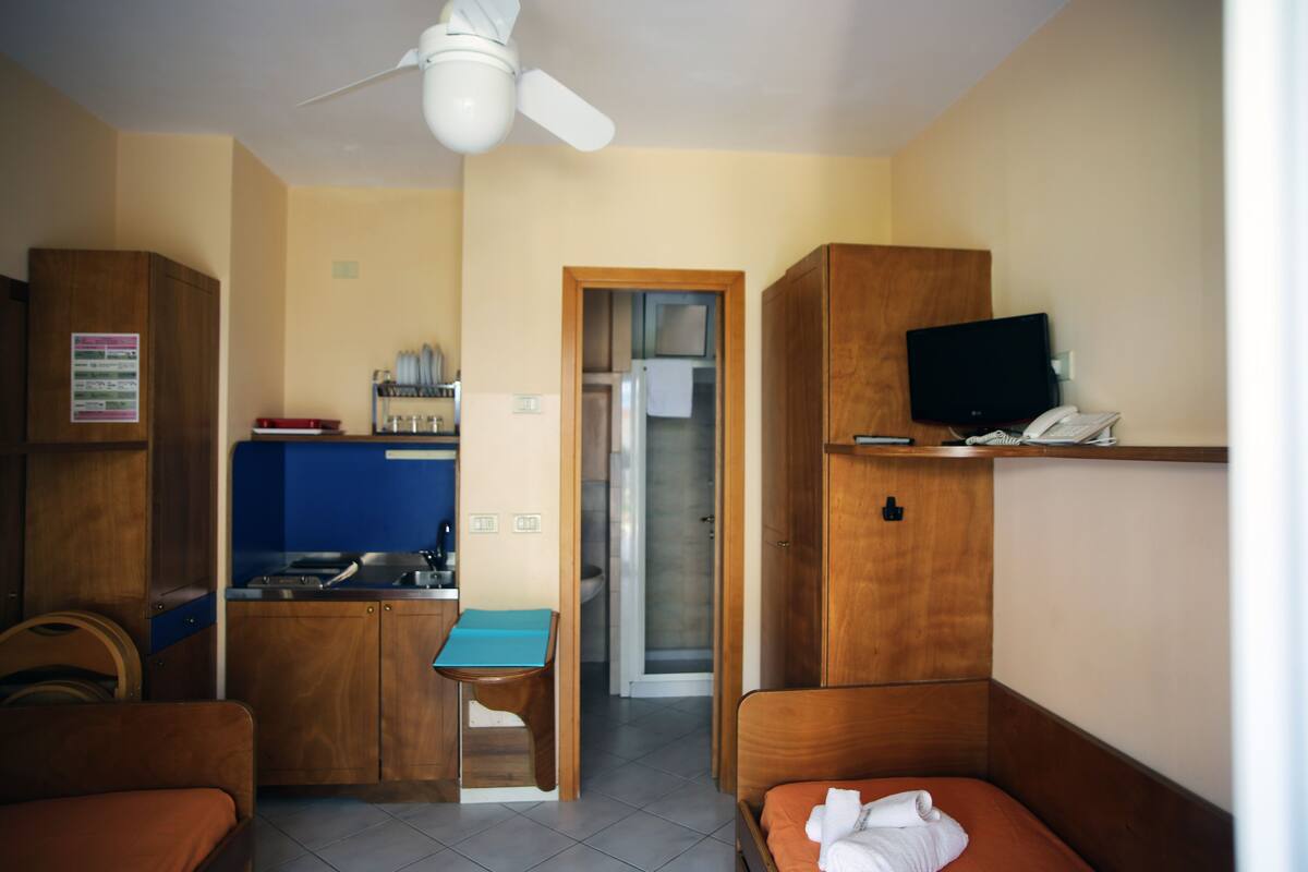 A compact hotel room with a kitchenette, a single bed, a small table, and a ceiling fan.