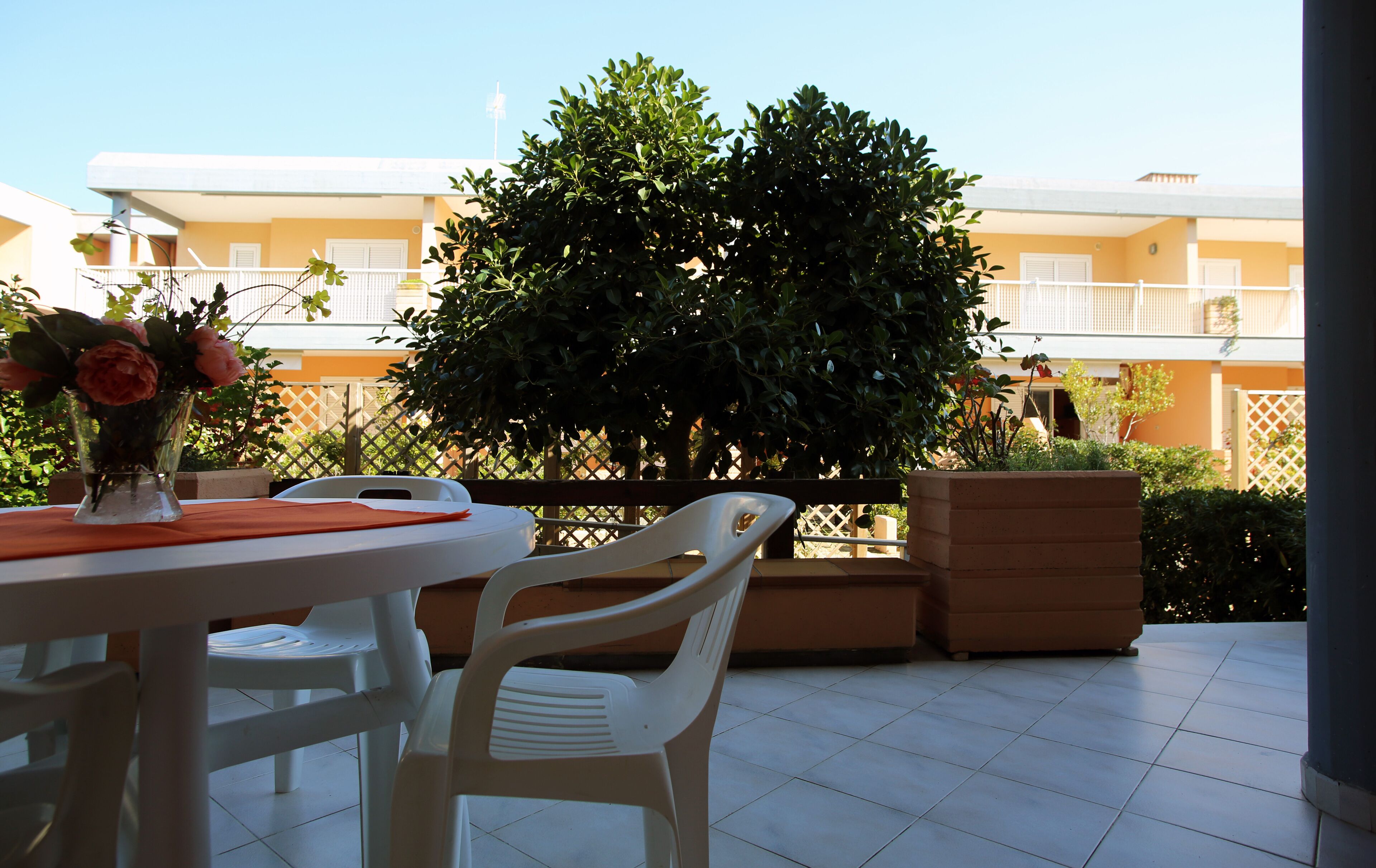 Un patio con una mesa y sillas, un florero con flores y un árbol grande.