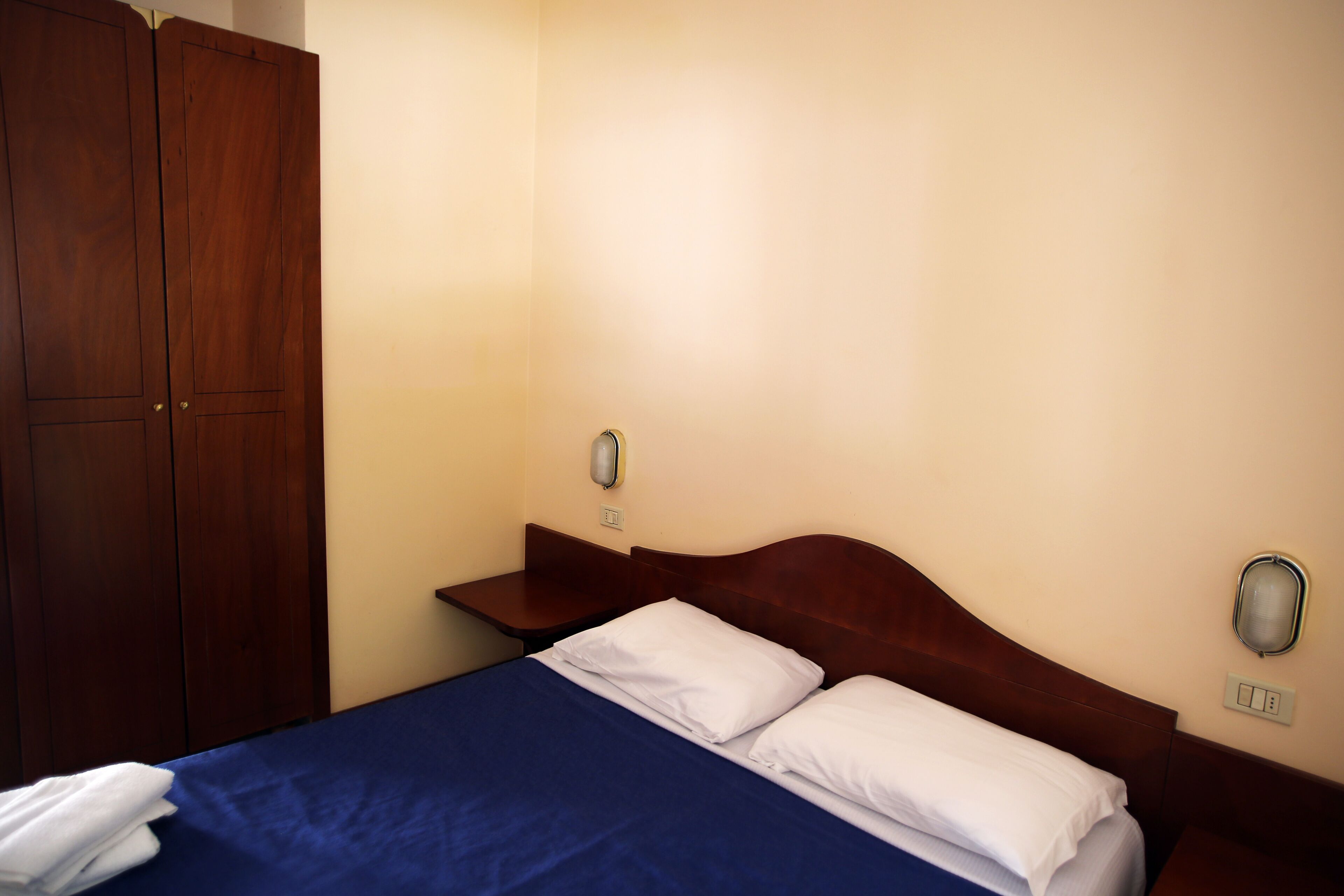 A hotel room with a bed, wooden wardrobe, bedside table, and wall-mounted light fixture.
