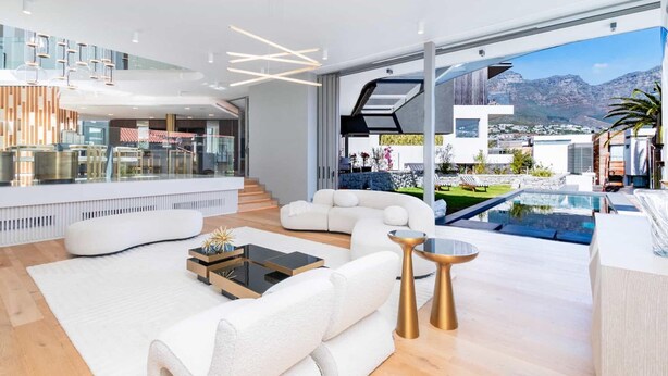 A modern living room with a white sofa, a coffee table, and a glass-top table. There is a pool visible through the large windows. - Grand Camps Bay 5bed w Views Pool Gym Villa Serena (Cape Town)