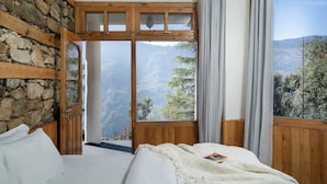 A bedroom with a stone wall, wooden beams, a large window with a view of mountains and trees, and a bed with white linens. - The Isisinga (Banjar)
