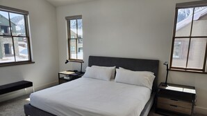 A bedroom with a large bed, two bedside tables, a bench, and a window with a view of a building and trees. - Snowcreek Resort Vacation Rental 1555 (Mammoth Lakes)