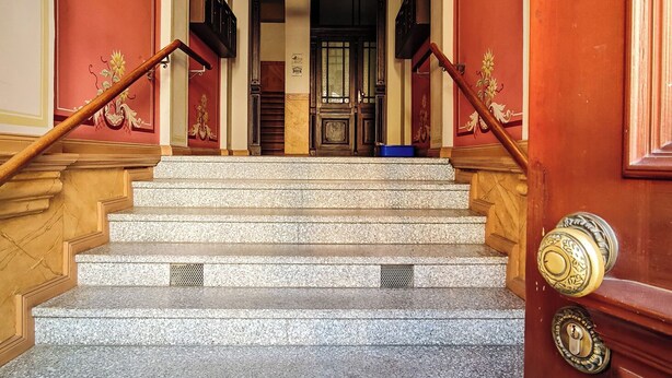 A grand staircase with marble steps leading to a dark wooden door, flanked by red walls and ornate decorations.
